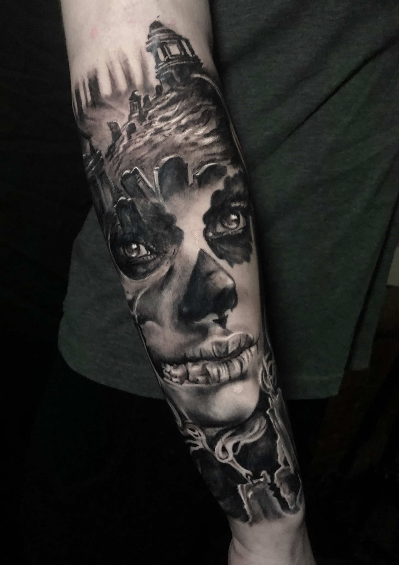 Black and gray forearm tattoo of a skull-faced woman with a graveyard and building in the background.