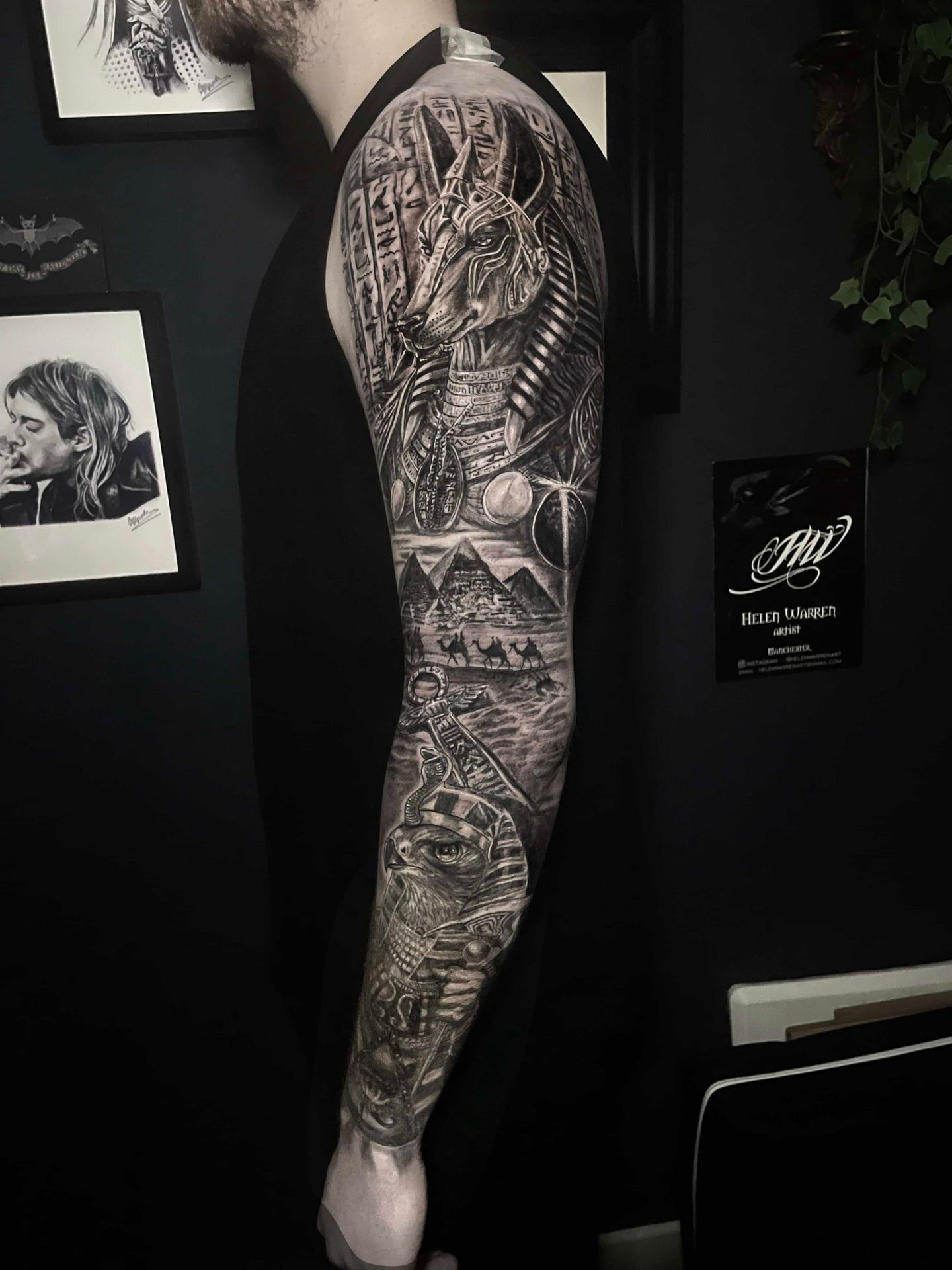 Man with detailed black and white arm sleeve tattoo. Dark background. Art on wall.