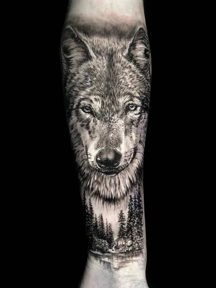 Detailed black and white wolf tattoo on an arm, with a forest landscape below the wolf's head.